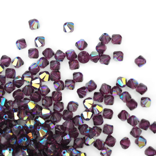 Austrian crystal 5328 bicone beads in the color Amethyst AB spilling out onto a white background.