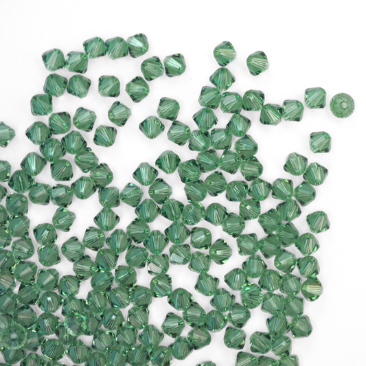 Austrian crystal 5328 bicone beads in the color Erinite spilling out onto a white background.