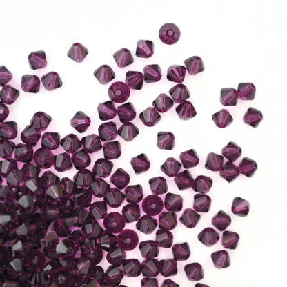 Austrian crystal 5328 bicone beads in the color Amethyst spilling out onto a white background. 