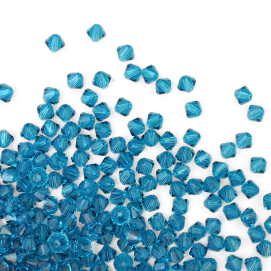 Austrian crystal 5328 bicone beads in the color Indicolite spilling out onto a white background. 