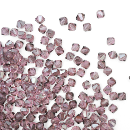 Austrian crystal 5328 bicone beads in the color Light Amethyst Satin spilling out onto a white background. 