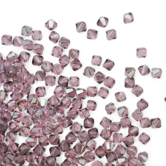 Austrian crystal 5328 bicone beads in the color Light Amethyst Satin spilling out onto a white background. 