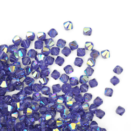 Austrian crystal 5328 bicone beads in the color Tanzanite AB spilling out onto a white background. 