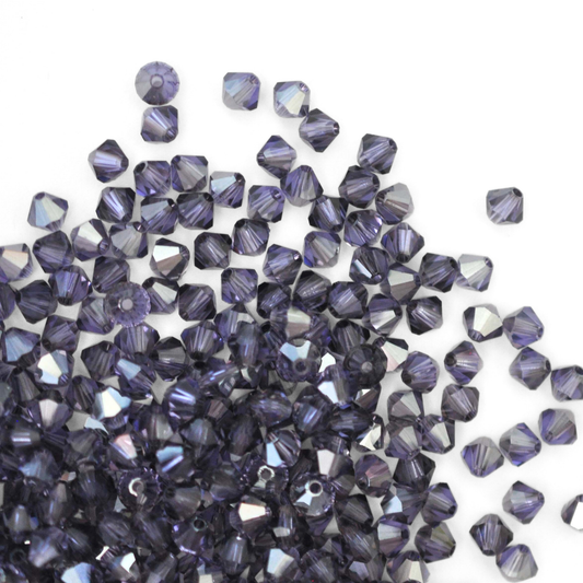 Austrian crystal 5328 bicone beads in the color Tanzanite Satin spilling out onto a white background. 