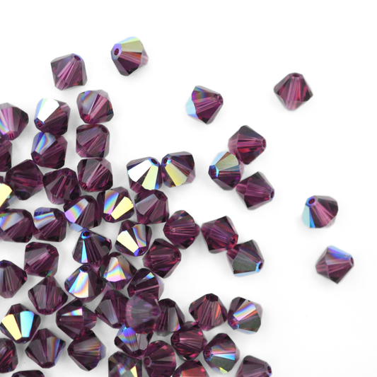 Austrian crystal 5328 bicone beads in the color Amethyst AB spilling out onto a white background. 