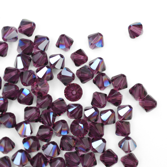 Austrian crystal 5328 bicone beads in the color Amethyst Moonlight spilling out onto a white background. 