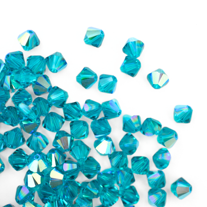 Austrian crystal 5328 bicone beads in the color Blue Zircon AB spilling out onto a white background. 