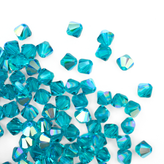 Austrian crystal 5328 bicone beads in the color Blue Zircon AB spilling out onto a white background. 
