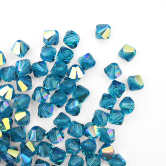 Austrian crystal 5328 bicone beads in the color Indicolite AB spilling out onto a white background. 