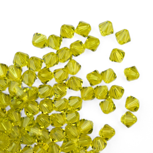 Austrian crystal 5328 bicone beads in the color Light Olivine spilling out onto a white background. 
