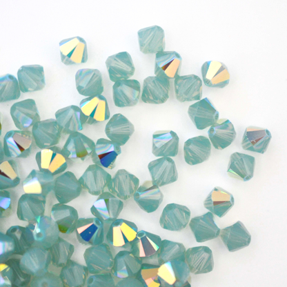 Austrian crystal 5328 bicone beads in the color Pacific Opal AB spilling out onto a white background. 