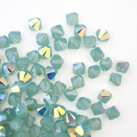 Austrian crystal 5328 bicone beads in the color Pacific Opal AB spilling out onto a white background. 