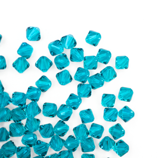 Austrian crystal 5328 bicone beads in the color Blue Zircon spilling out onto a white background. 